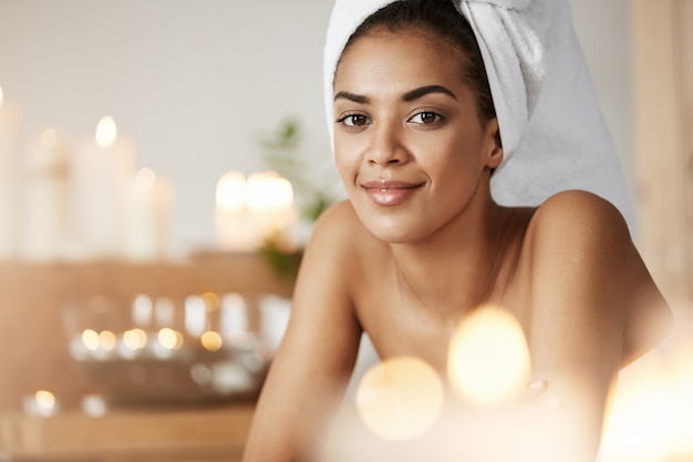 Retrato da mulher africana bonita com a toalha na cabeça que sorri descansando no salão spa.