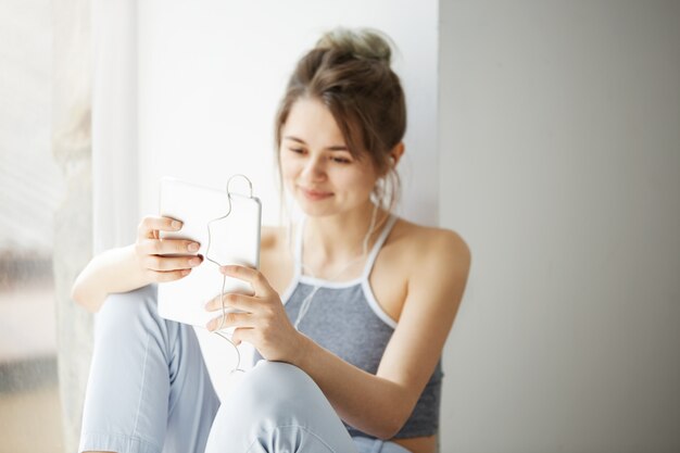 Retrato da mulher alegre adolescente nova nos fones de ouvido que sorri olhando a Web surfando da navigação da tabuleta que senta-se perto da janela sobre a parede branca.