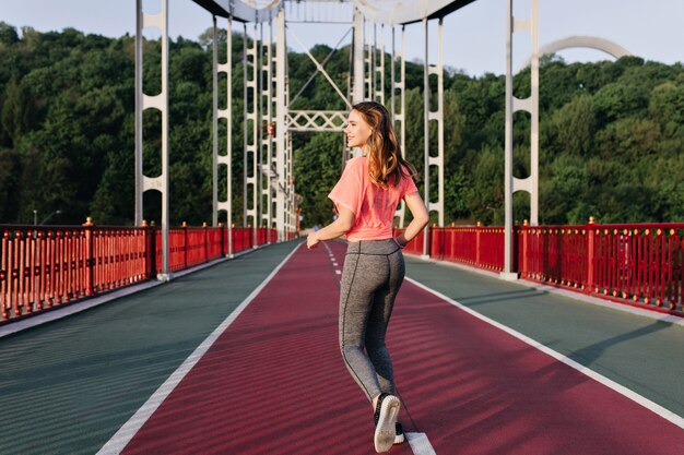 Retrato da parte de trás da menina alegre correndo pela manhã e apreciando a vista da natureza. Foto ao ar livre de senhora inspirada fazendo exercício.