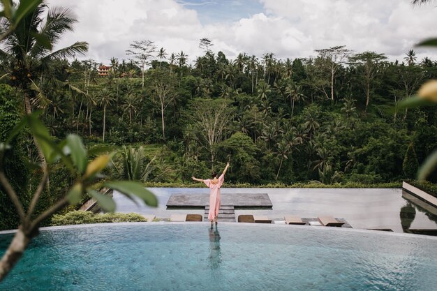 Retrato da parte de trás do modelo feminino em vestido rosa, olhando para a floresta tropical. Tiro ao ar livre de mulher graciosa dançando perto da piscina.