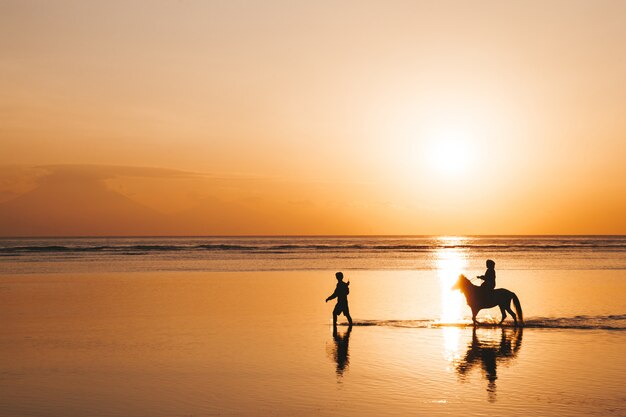 Retrato da silhueta do jovem casal romântico andando a cavalo na praia. Menina e o namorado ao pôr do sol colorido dourado