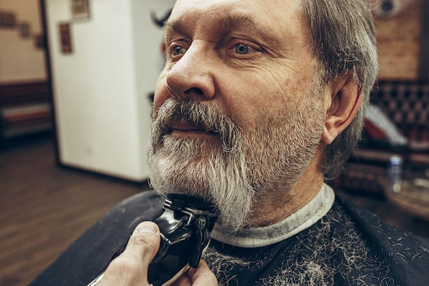 Retrato da vista lateral do close-up do homem caucasiano barbudo sênior considerável que obtém a barba que prepara na barbearia moderna.