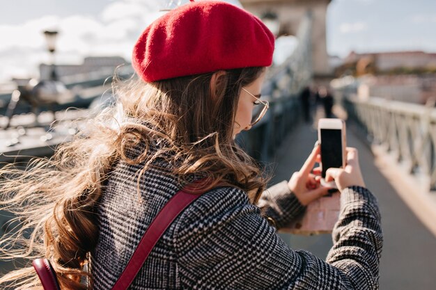 Retrato das costas de uma mulher elegante em uma jaqueta de tweed segurando o telefone e tirando uma foto da paisagem da cidade