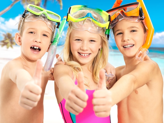 Retrato das crianças felizes com gesto de polegar para cima na praia. crianças em idade escolar juntos em trajes de banho de cor brilhante com máscara de natação na cabeça.