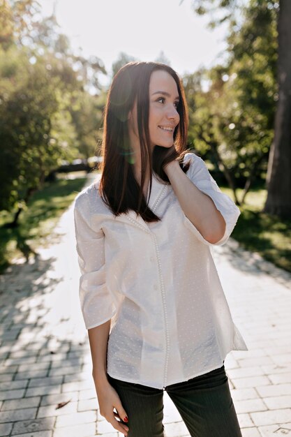 Retrato de adorável garota agitada com cabelo escuro e vestindo blusa branca e jeans está sorrindo de lado no parque verde ensolarado