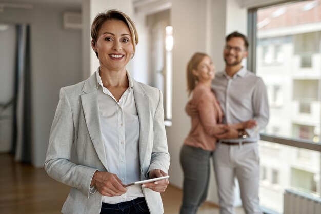 Retrato de agente imobiliário feminino olhando para a câmera enquanto casal feliz em pé no fundo