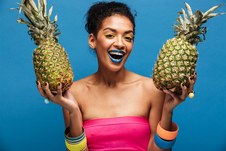 Foto grátis retrato de alegre mulher afro-americana com maquiagem moda sorrindo e segurando dois abacaxis em ambas as mãos isoladas, sobre parede azul