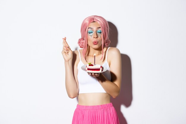 Retrato de aniversariante esperançoso na peruca rosa, fazendo desejo com os dedos cruzados, segurando o bolo de aniversário, em pé.