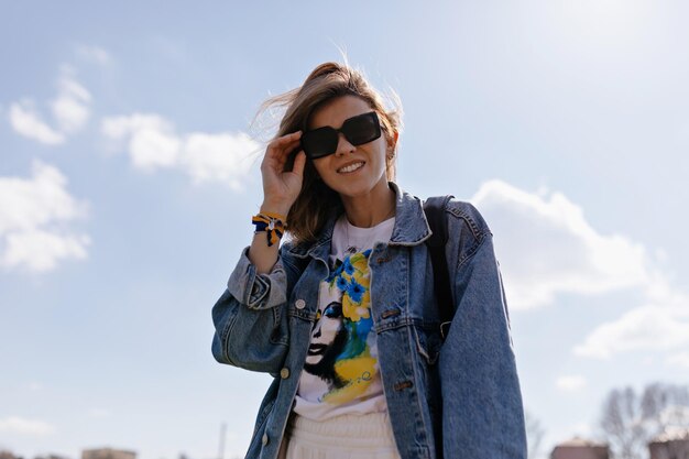 Retrato de baixo da menina feliz sorridente em óculos de sol e jaqueta jeans e camiseta com estampa azul e amarela está tocando óculos e sorrindo para a câmera contra o céu azul