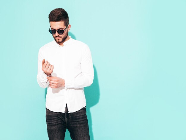 Retrato de bonito modelo confiante Homem elegante sexy vestido com camisa branca e jeans Moda hipster masculino posando perto da parede azul no estúdio isolado em óculos de sol