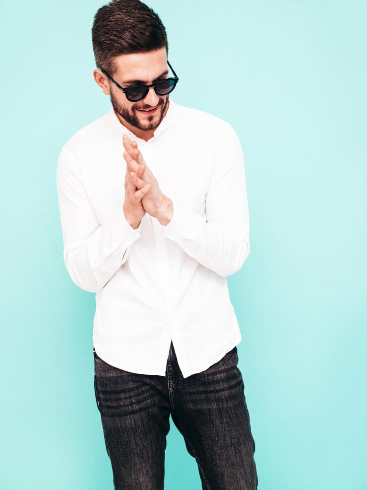 Foto grátis retrato de bonito modelo confiante homem elegante sexy vestido com camisa branca e jeans moda hipster masculino posando perto da parede azul no estúdio isolado em óculos de sol