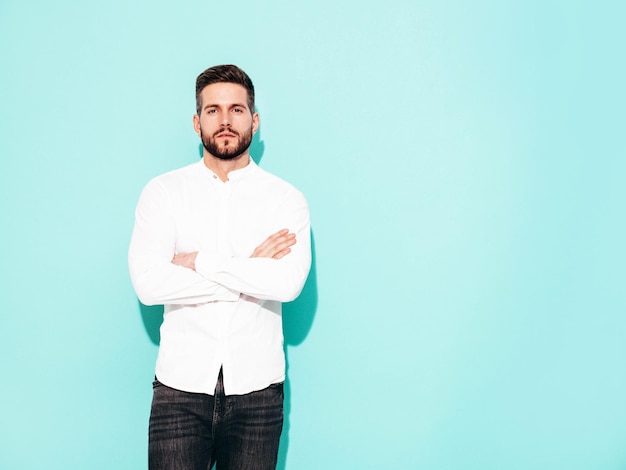 Retrato de bonito modelo confiante Homem elegante sexy vestido de camisa branca e jeans Moda hipster masculino posando perto da parede azul no estúdio isolado