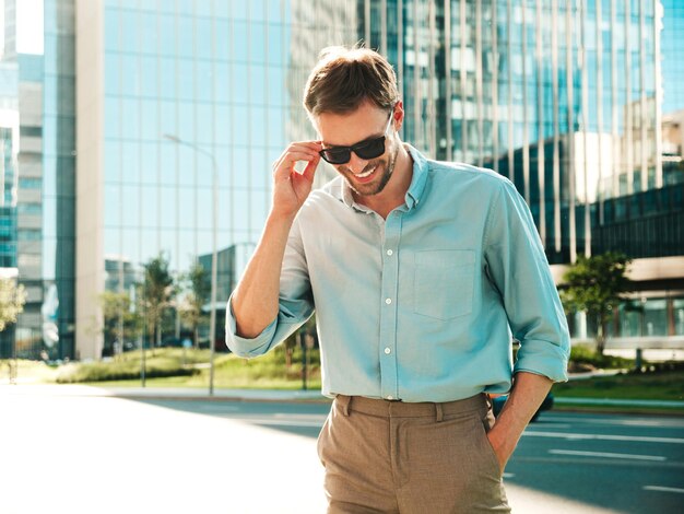 Retrato de bonito sorridente elegante hipster lambersexual modelo Homem moderno vestido com camisa azul Moda masculina posando no fundo da rua perto de arranha-céus em óculos de sol