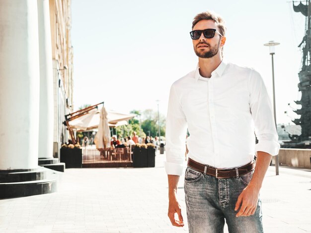 Retrato de bonito sorridente elegante hipster lambersexual modelo Homem moderno vestido com camisa branca Moda masculina posando no fundo da rua em óculos de sol Ao ar livre ao pôr do sol