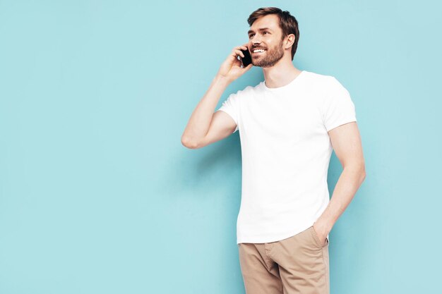Retrato de bonito sorridente elegante hipster lambersexual modelo Homem sexy vestido de camiseta e jeans Moda masculina isolada na parede azul no estúdio falando no smartphone com amigos