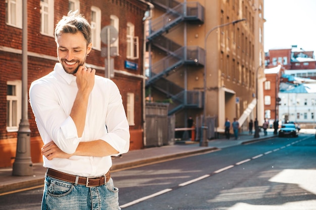 Retrato de bonito sorridente hipster elegante modelo lambersexual Homem moderno vestido de camisa branca Moda pensativo masculino posando no fundo da rua Ao ar livre ao pôr do sol