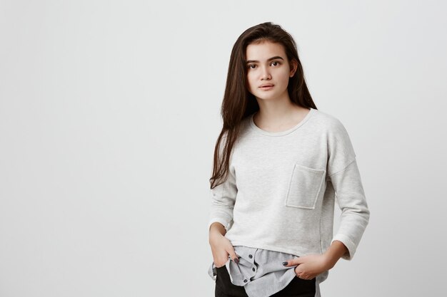 Retrato de cabelo de mulher adolescente bonita com rosto oval, olhos atraentes escuros e cabelo longo e reto, vestido casualmente, sentindo-se relaxado em pé com as mãos nos bolsos, posando dentro de casa