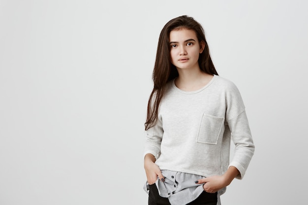 Retrato de cabelo de mulher adolescente bonita com rosto oval, olhos atraentes escuros e cabelo longo e reto, vestido casualmente, sentindo-se relaxado em pé com as mãos nos bolsos, posando dentro de casa