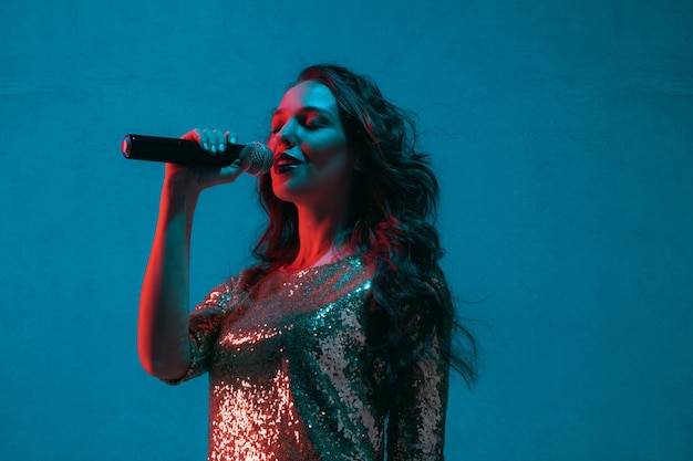 Foto grátis retrato de cantora caucasiana isolado no fundo azul do estúdio em luz de néon. bela modelo feminino em um vestido brilhante com microfone. conceito de emoções humanas, expressão facial, anúncio, música, arte.