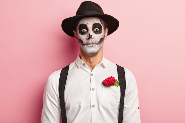 Retrato de cara bonito assustador feito maquiagem para evento de Halloween, tem imagem de vampiro ou fantasma, flor de rosa vermelha no bolso da camisa branca, usa chapéu preto, tem aparência assustadora