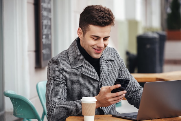 Retrato de cara de sucesso descansando no café de rua, trabalhando com o notebook e digitando a mensagem de texto em seu telefone móvel