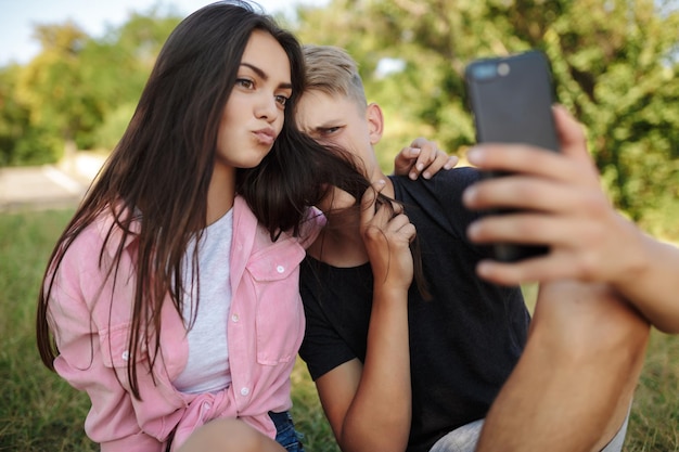 Retrato de casal fofo sentado no gramado e se abraçando enquanto fazia selfie engraçada no parque. Lindo casal tirando fotos na câmera frontal do celular