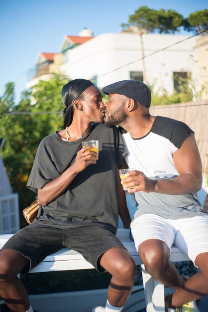 Retrato de casal gay africano feliz beijando no banco. Dois homens em roupas casuais, sentado no banco com copos de suco, beijando os lábios um do outro. Amor homossexual e relações do conceito de casais LGBT