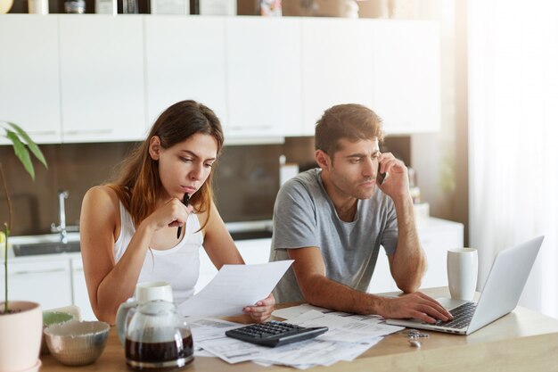 Retrato de casal jovem: fêmea lendo atentamente o documento e macho sentado na frente do laptop aberto e conversando com o parceiro de negócios por telefone inteligente, estar ocupado com a elaboração de relatório financeiro