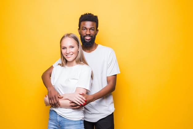 Retrato de casal romântico multirracial se abraçando, posando juntos e sorrindo, em pé sobre a parede amarela