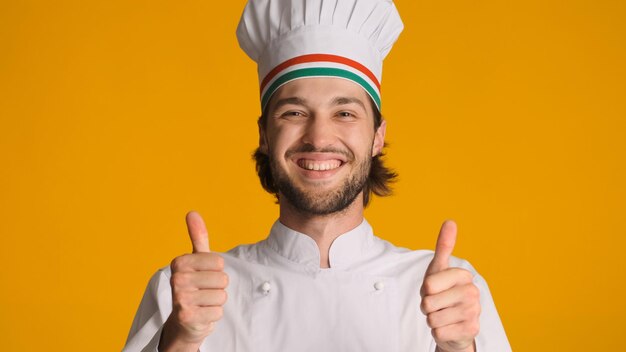 Retrato de chef masculino feliz de uniforme, mantendo os polegares para cima na câmera contra um fundo colorido Homem bonito vestido com chapéu de chef mostrando gesto aprovado