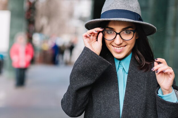 Retrato de cidade elegante encantadora jovem com casaco cinza, chapéu, andar na rua. Óculos pretos modernos, sorrindo, expressando verdadeiras emoções felizes, estilo de vida luxuoso, modelo elegante.