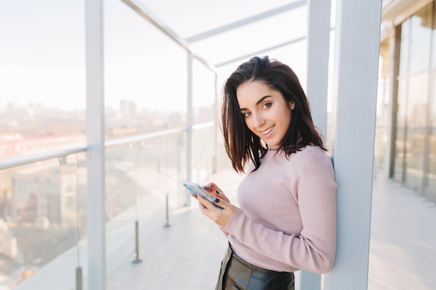 Retrato de cidade elegante jovem morena elegante usando telefone no terraço na vista da cidade. Mulher de negócios atraente, bom humor, sorrindo.