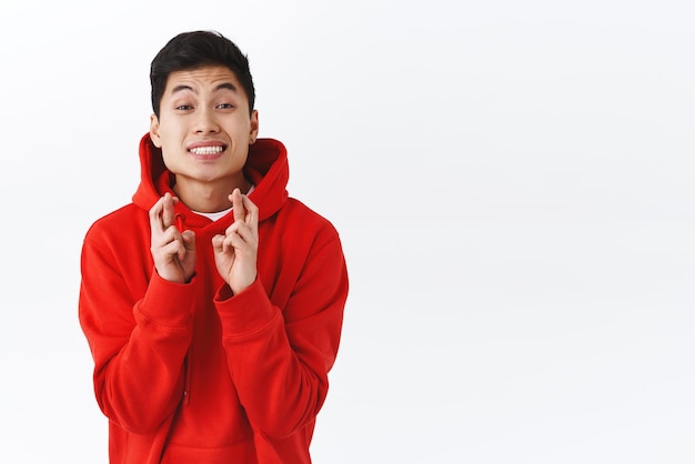 Foto grátis retrato de cintura de homem asiático entusiasmado atraente com capuz vermelho implorando para que o sonho se torne realidade sorrindo otimista dedos cruzados boa sorte fazer desejo ou rezar fundo branco