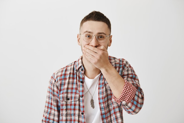 Foto grátis retrato de cintura de homem encantador alegre de óculos e com corte de cabelo na moda cobrindo a boca tentando segurar a risada em pé contra um fundo cinza guy colocou aparelho nos dentes e tímido para mostrá-lo