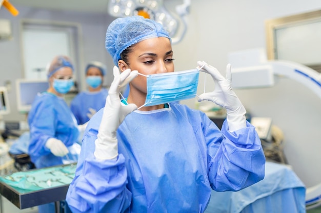 Retrato de cirurgiã usando máscara cirúrgica e boné Médico qualificado está trabalhando na sala de cirurgia iluminada Ela está no hospital