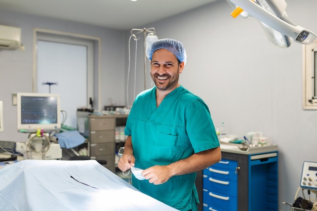 Retrato de cirurgião masculino no teatro de operação, olhando para a câmera mostrando o gesto OK Doutor em jaleco e máscara médica na moderna sala de cirurgia do hospital