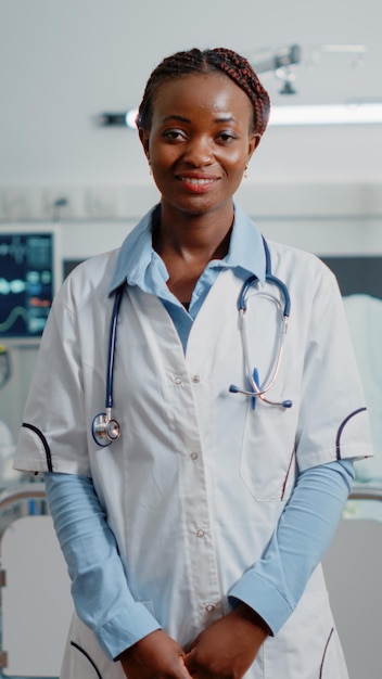 Retrato de clínico geral em pé na enfermaria do hospital, pronto para tratar o paciente com doença. Médico da mulher com estetoscópio e jaleco branco trabalhando com equipamentos médicos na clínica.