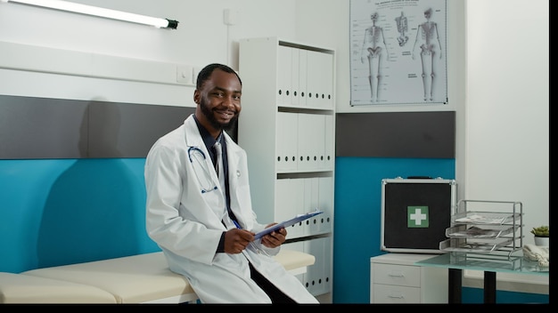 Foto grátis retrato de clínico geral tomando notas no consultório médico, esperando para iniciar o exame de check-up com o paciente. médico masculino com jaleco branco usando documentos de consulta no consultório da clínica.