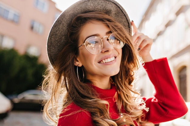 Retrato de close-up de alegre mulher branca de óculos tocando seu chapéu em desfocar o fundo