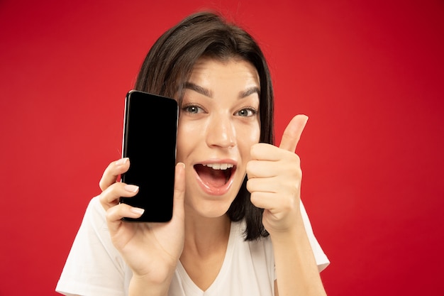 Retrato de close up de mulher jovem caucasiana no fundo vermelho do estúdio. Bela modelo feminino em camisa branca. Conceito de emoções humanas, expressão facial, vendas. Segurando o telefone, parece feliz, mostra-se bem.