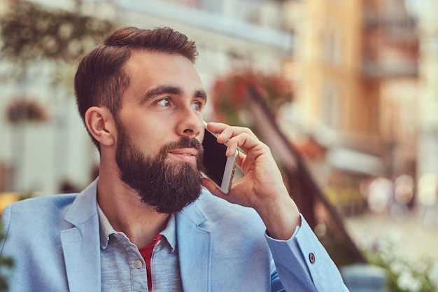 Retrato de close-up de um empresário barbudo elegante com um corte de cabelo elegante, falando por telefone, bebe um copo de suco fresco, sentado em um café ao ar livre.