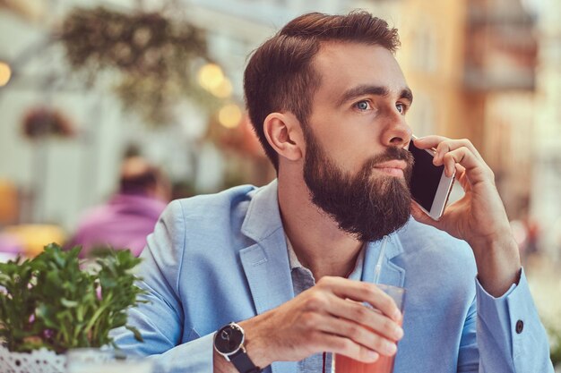 Retrato de close-up de um empresário barbudo elegante com um corte de cabelo elegante, falando por telefone, bebe um copo de suco fresco, sentado em um café ao ar livre.