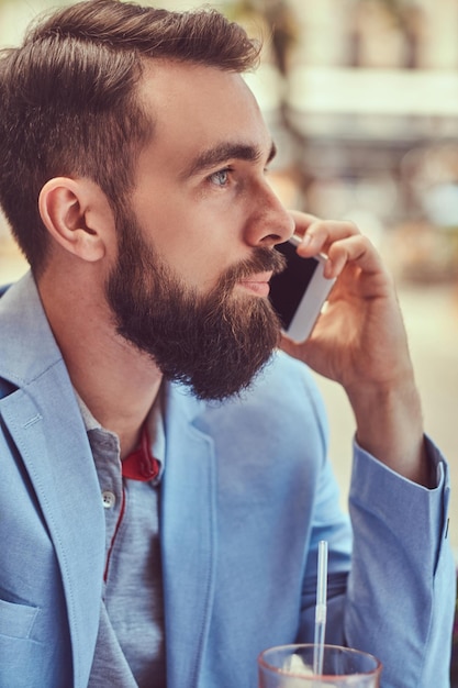 Foto grátis retrato de close-up de um empresário barbudo elegante com um corte de cabelo elegante, falando por telefone, bebe um copo de suco fresco, sentado em um café ao ar livre.