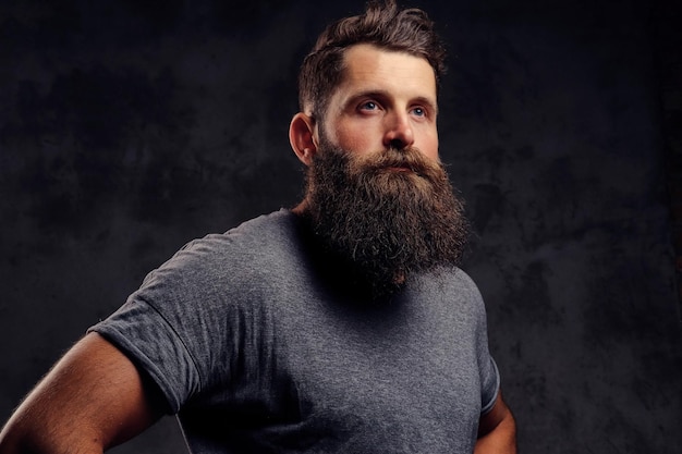 Retrato de close-up de um hipster com barba cheia e corte de cabelo elegante, vestido com uma camiseta cinza, fica em um estúdio em um fundo escuro.