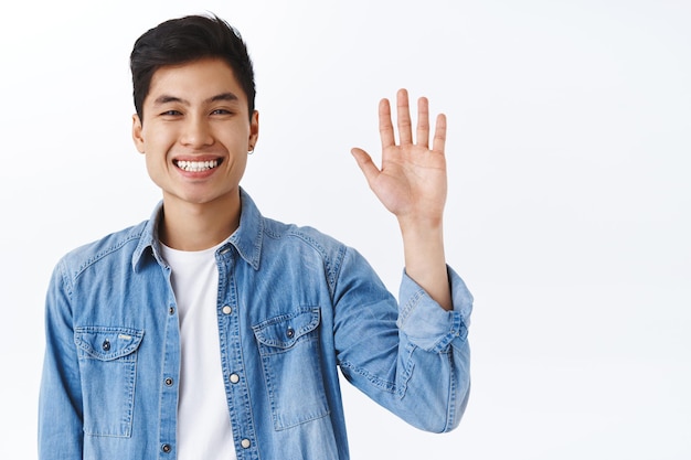 Retrato de close-up de um homem asiático atraente e amigável acenando para dizer oi, gesto informal de saudação, rindo e sorrindo ao dar as boas-vindas aos novos membros.
