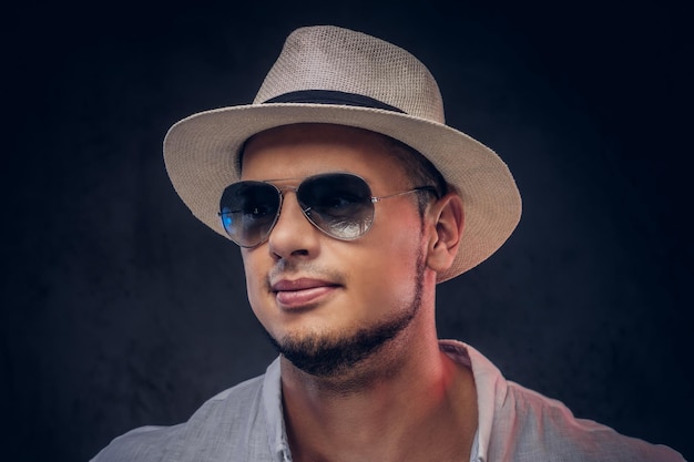 Retrato de close-up de um homem barbudo elegante bonito em uma camisa branca, chapéu panamá e óculos de sol. Isolado em fundo escuro.