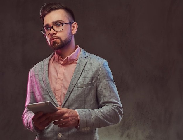 Retrato de close-up de um homem barbudo elegante confiante com penteado e óculos de sol em um terno cinza e camisa rosa, detém um tablet em um estúdio. Isolado em um fundo cinza.