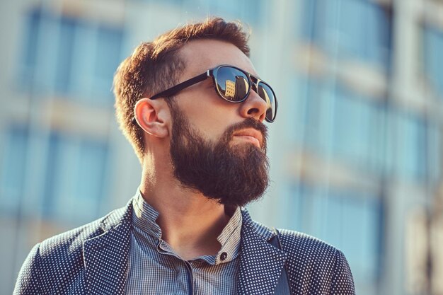 Retrato de close-up de um homem barbudo vestindo roupas casuais e óculos de sol, de pé em uma rua da cidade contra um arranha-céu.