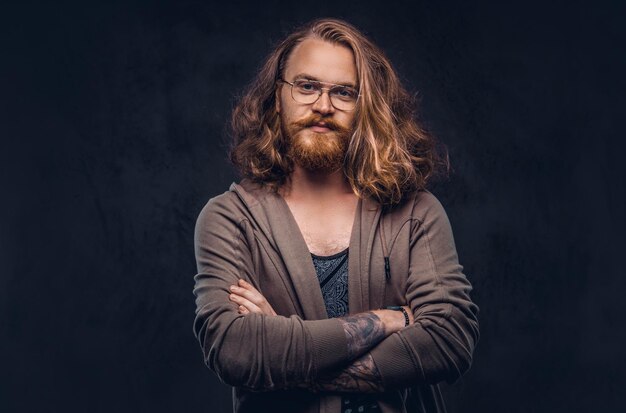 Retrato de close-up de um homem ruiva hipster com cabelos longos e exuberantes e barba cheia, vestido com roupas casuais em pé em um estúdio, olhando para longe. Isolado em um fundo escuro.