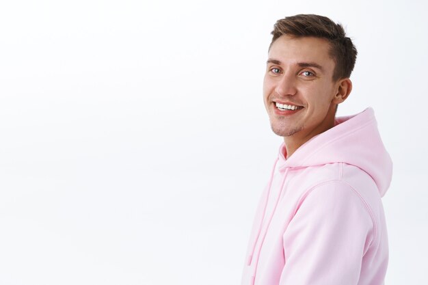 Retrato de close-up de um jovem estudante bonito com cabelo loiro, eriçado, em pé de perfil e virado para a câmera com um sorriso radiante, parecendo satisfeito feliz, parede branca de pé